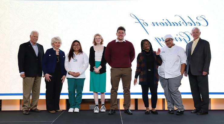 The scholarship recipients and donors standing in a line in front of a large screen that says Celebration of Giving in a blue cursive font