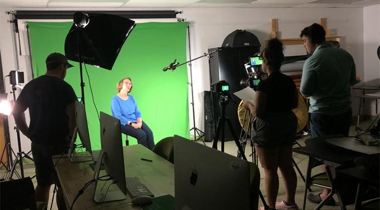 Three Visual Communication students in the foreground standing amidst filming equipment. They are filming Bobbie Hemmings who is sitting on a stool in front of a green screen.